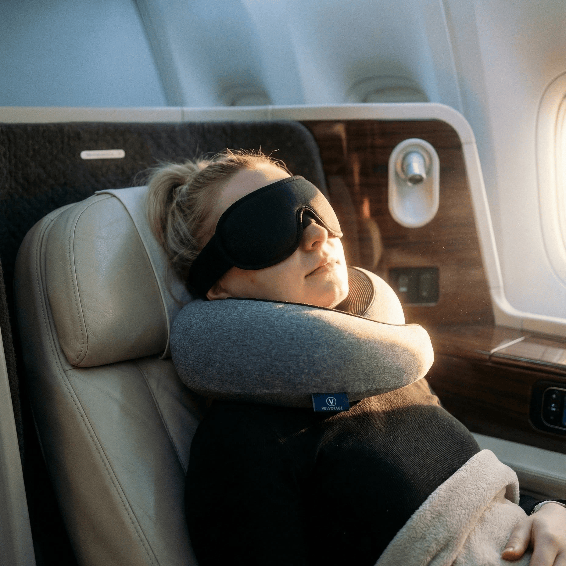 Person sleeping on an airplane with a neck pillow and eye mask.